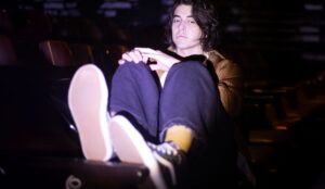 Cantor Lean sentado entre as poltronas de um teatro, usando jaqueta marrom, calça preta e tênis, em foto com iluminação baixa que destaca a estética indie do artista.