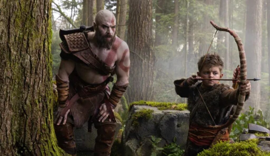 Kratos e Atreus aparecem em uma floresta em imagem promocional da série de God of War. Enquanto Kratos observa o ambiente, Atreus mira um arco e flecha.