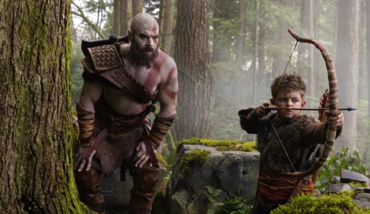 Kratos e Atreus aparecem em uma floresta em imagem promocional da série de God of War. Enquanto Kratos observa o ambiente, Atreus mira um arco e flecha.
